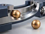 Konzeptionelle CGI-Darstellung: warme bronzefarbene Kugel auf einer glatten gekrümmten Stahlschiene sowie eine davon getrennte intakte Kugel; veranschaulicht ein Kontaktpaar aus verschiedenen Werkstoffen statt abstrakter Härteaussagen