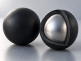 CGI conceptuelle : coupe d’une sphère à revêtement noir en caoutchouc révélant un noyau métallique argenté solide entouré d’une épaisse coque d’élastomère noir uniforme, à côté d’une sphère intacte correspondante ; adhérence du revêtement représentée simplement, sans écailles éclatées