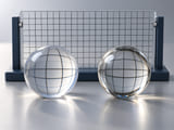 CGI conceptual: esfera acrílica transparente y esfera de vidrio transparente junto a una cuadrícula sencilla de observación óptica; refracciones diferentes, físicamente plausibles; sin etiquetas ni afirmaciones de medición